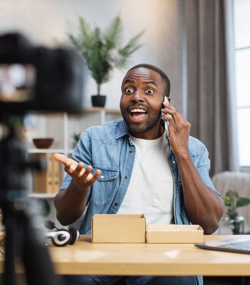 Happy african american man testing new modern smartphone while recording video on camera at home. Male influencer doing live stream. People and social networks concept.